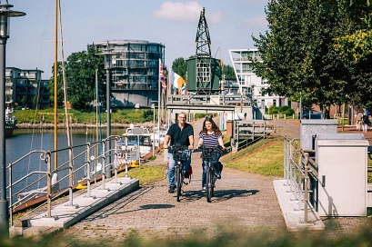 Zwei Menschen fahren bei Sonnenschein nebeneinander auf Fahrrädern am Stader Hafen entlang. Im Hintergrund sind Boote, moderne Gebäude und ein alter Kran zu sehen.