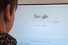 Symbolbild - Google Eine Person mit kurzen braunen Haaren blickt von der Seite auf einen großen Bildschirm mit der Google-Startseite. Die Person trägt einen silberfarbenen Ohrring sowie ein schwarz-beige gemustertes Oberteil. Der helle Bildschirm dominiert das Bild, darauf ist mittig das farbige Google-Logo auf weißem Hintergrund zu sehen. Die Aufnahme wirkt ruhig und konzentriert und vermittelt den Eindruck einer Internetsuche oder digitalen Recherche.