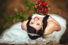 Symbolbild - Hochzeit Eheschließung Eine lächelnde Braut blickt von unten in die Kamera und hält einen Strauß roter Rosen in der Hand. Sie trägt ein weißes Brautkleid, eine zarte Tiara im dunklen Haar und ist von einem natürlichen, grünen Hintergrund umgeben.