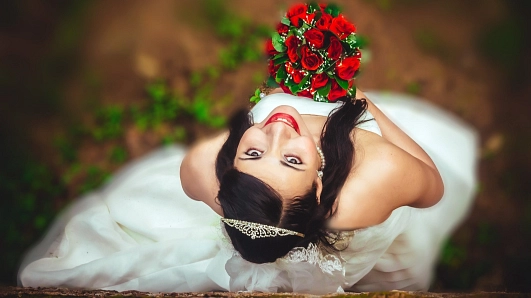 Eine lächelnde Braut blickt von unten in die Kamera und hält einen Strauß roter Rosen in der Hand. Sie trägt ein weißes Brautkleid, eine zarte Tiara im dunklen Haar und ist von einem natürlichen, grünen Hintergrund umgeben.