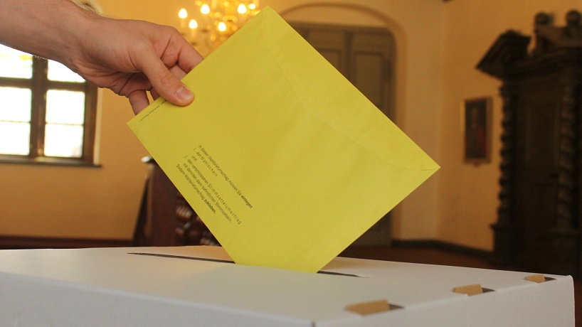 Zu sehen ist die Detailaufnahme einer Hand, die einen Wahlumschlag in eine Wahlurne einwirft