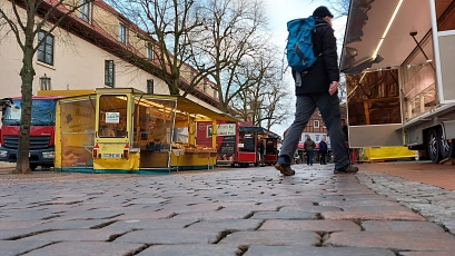Symbolbild-Wochenmarkt2.jpg Blick auf den Stader Wochenmarkt: Verschiedene Verkaufsstände mit frischen Lebensmitteln reihen sich entlang der gepflasterten Straße. Im Vordergrund läuft eine Person mit blauem Rucksack vorbei, im Hintergrund sind weitere Marktbesucher*innen und historische Gebäude zu sehen. Die Szene wirkt lebendig und einladend. © Hansestadt Stade
