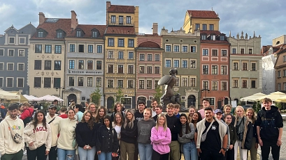Eine Gruppe junger Menschen steht dicht nebeneinander auf einem historischen Platz in Warschau, Polen. Im Hintergrund sind bunte, detailreich verzierte Altstadthäuser in Gelb, Grün, Rosa, Grau und Ocker mit roten Ziegeldächern zu sehen. In der Mitte des Platzes befindet sich eine bronzene Meerjungfrauenstatue mit Schild und Schwert – das Wahrzeichen der Stadt. Die Jugendlichen tragen überwiegend legere Kleidung in gedeckten Farben wie Beige, Grau, Schwarz oder Rosa. Einige lächeln, andere blicken neutral in die Kamera. Der Himmel ist teils bewölkt, die Stimmung wirkt lebendig und freundlich.