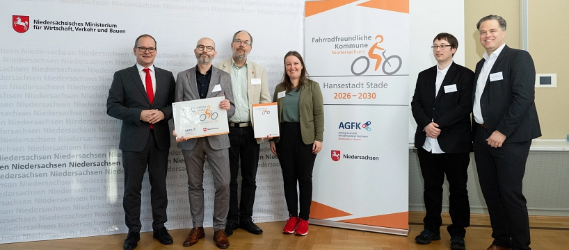Sechs Personen stehen nebeneinander in einem festlich beleuchteten Raum auf hellem Parkettboden. Im Zentrum halten zwei Männer Urkunden in der Hand, die die Auszeichnung „Fahrradfreundliche Kommune Niedersachsen“ zeigen. Links steht ein Mann im grauen Anzug mit roter Krawatte, rechts daneben zwei Männer in grauen Anzügen mit offenen Hemden – einer mit Brille und Bart. Eine junge Frau mit langen braunen Haaren trägt ein olivgrünes Oberteil, schwarzen Blazer, schwarze Hose und auffällige rote Turnschuhe. Rechts daneben stehen zwei Männer in dunklen Anzügen; einer trägt ein weißes Hemd offen über der Hose. Die Stimmung ist freundlich und professionell. Im Hintergrund stehen zwei weiße Roll-Ups: links mit dem Logo des Niedersächsischen Ministeriums für Wirtschaft, Verkehr und Bauen, rechts mit dem Logo der Auszeichnung für die Hansestadt Stade für den Zeitraum 2026 bis 2030.