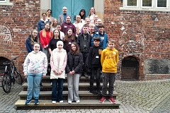 Eine größere Gruppe von rund 25 Kindern und Jugendlichen sowie erwachsenen Begleitpersonen steht auf breiten Steinstufen vor einem historischen Backsteingebäude. Die Fassade ist aus rötlichen Ziegeln gemauert, mit sichtbaren Bögen und zwei kleinen, vergitterten Öffnungen im unteren Bereich. Im Hintergrund befindet sich eine große, türkisfarbene Doppeltür, darüber helle Fensterrahmen. Die Gruppe ist in mehreren Reihen angeordnet und blickt in die Kamera. Die Stimmung wirkt freundlich und ruhig, mit leicht ernsten, aber offenen Gesichtsausdrücken. Die Kinder und Jugendlichen tragen überwiegend legere Alltagskleidung in unterschiedlichen Farben, darunter Jacken, Hoodies und Pullover in Schwarz, Grau, Blau, Rot und Gelb. Einzelne Kleidungsstücke setzen auffällige Farbakzente, etwa ein leuchtend gelber Kapuzenpullover oder eine rote Sweatjacke. Eine Person trägt ein helles Kopftuch. Die beiden erwachsenen Personen stehen in der hinteren Reihe, etwas erhöht. Sie sind schlichter und etwas formeller gekleidet, unter anderem mit einem grauen Sakko. Links neben der Treppe steht ein schwarzes Fahrrad. Der Boden im Vordergrund besteht aus grauem Kopfsteinpflaster. Insgesamt vermittelt das Bild den Eindruck eines Gruppenfotos im Rahmen eines gemeinsamen Besuchs oder Projekts.